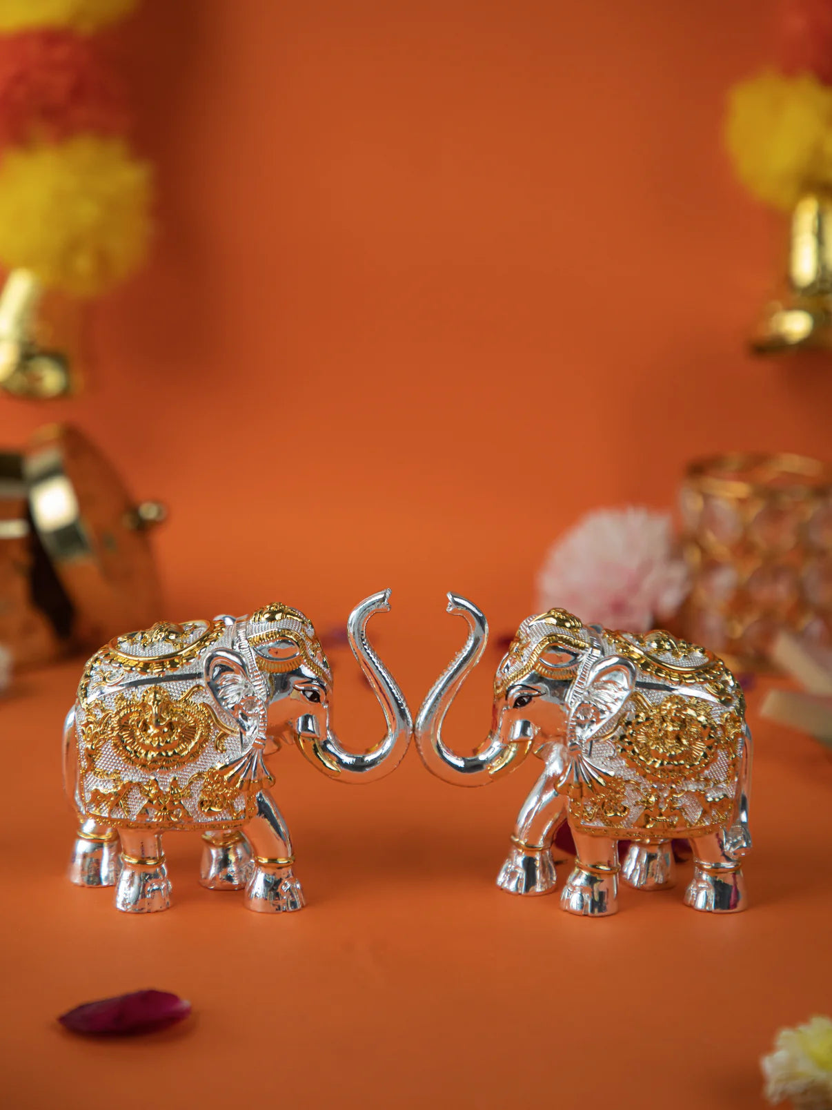 close-up shot of a pair of gold and silver elephant statues with their trunks intertwined. The elephants are intricately decorated with detailed patterns. The image is set against an orange background, with blurred flowers and other decorative items visible in the background.