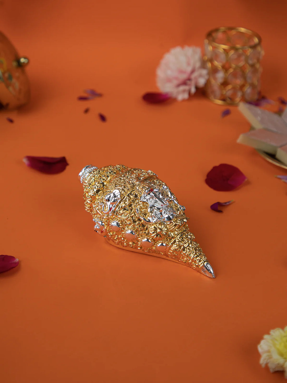 A golden and silver conch shell, or shankh, with intricate carvings rests on a vibrant orange surface, surrounded by scattered pink and yellow flower petals. In the background, there is a golden decorative object and some sweets.