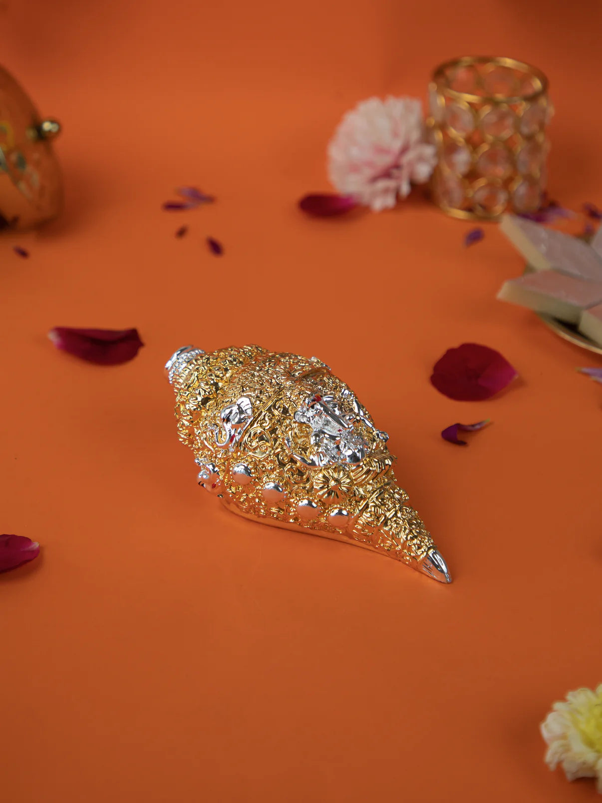 A golden and silver conch shell, or shankh, with intricate carvings rests on a vibrant orange surface, surrounded by scattered pink and yellow flower petals. In the background, there is a golden decorative object and some sweets.