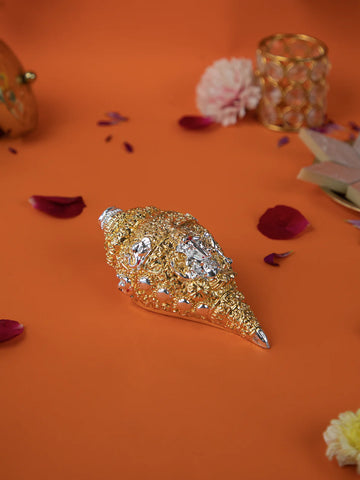 A golden and silver conch shell, or shankh, with intricate carvings rests on a vibrant orange surface, surrounded by scattered pink and yellow flower petals. In the background, there is a golden decorative object and some sweets.
