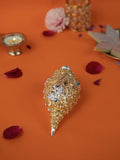 A detailed golden conch shell, known as a Shankh, with silver accents and an engraved image of Ganesha, is placed on an orange surface. Surrounding the Shankh are rose petals, a lit diya (oil lamp), and a plate of sweets. The background is a mix of orange and black, with a decorative object visible in the upper right corner.