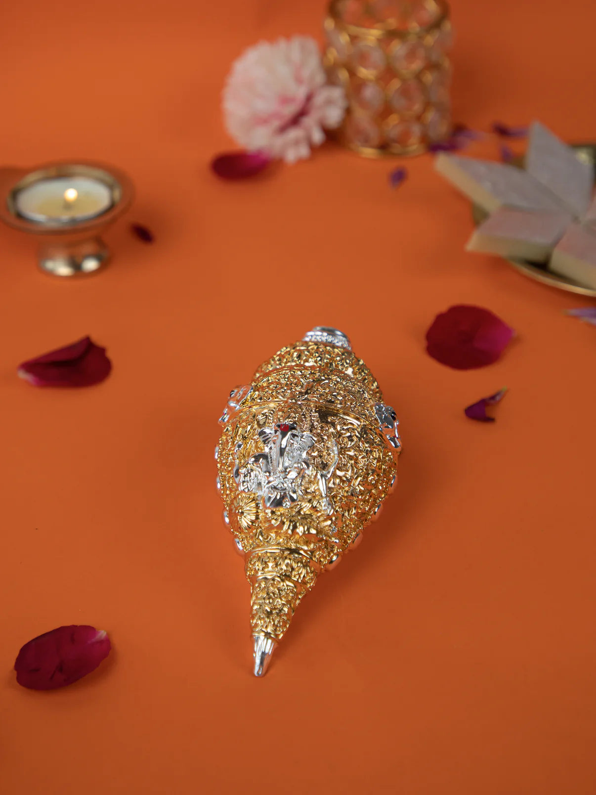 A detailed golden conch shell, known as a Shankh, with silver accents and an engraved image of Ganesha, is placed on an orange surface. Surrounding the Shankh are rose petals, a lit diya (oil lamp), and a plate of sweets. The background is a mix of orange and black, with a decorative object visible in the upper right corner.