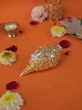 A detailed, gold-plated shankh (conch shell) featuring a Ganesha figure, surrounded by flowers and a lit diya, against an orange background. The shankh is a Hindu sacred object, symbolizing blessings and prosperity.