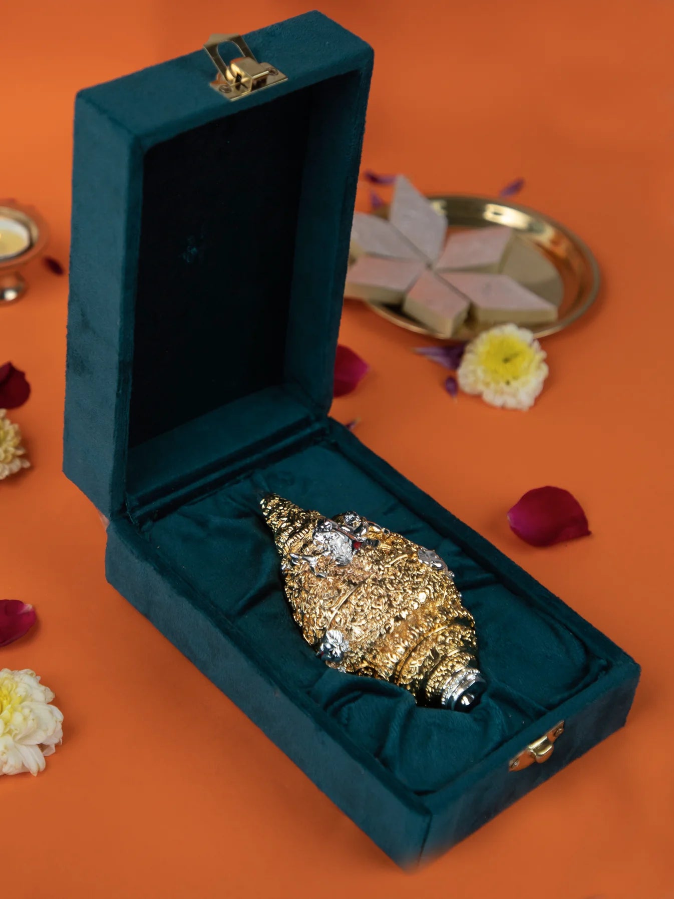 An image of a golden conch shell, also known as a shankh, decorated with silver and jewels, is displayed in an open teal velvet gift box. In the background, there is a silver tray with a type of Indian sweet called kaju katli, along with some flower petals. The item is often used for religious purposes, home decor, or as a gift.