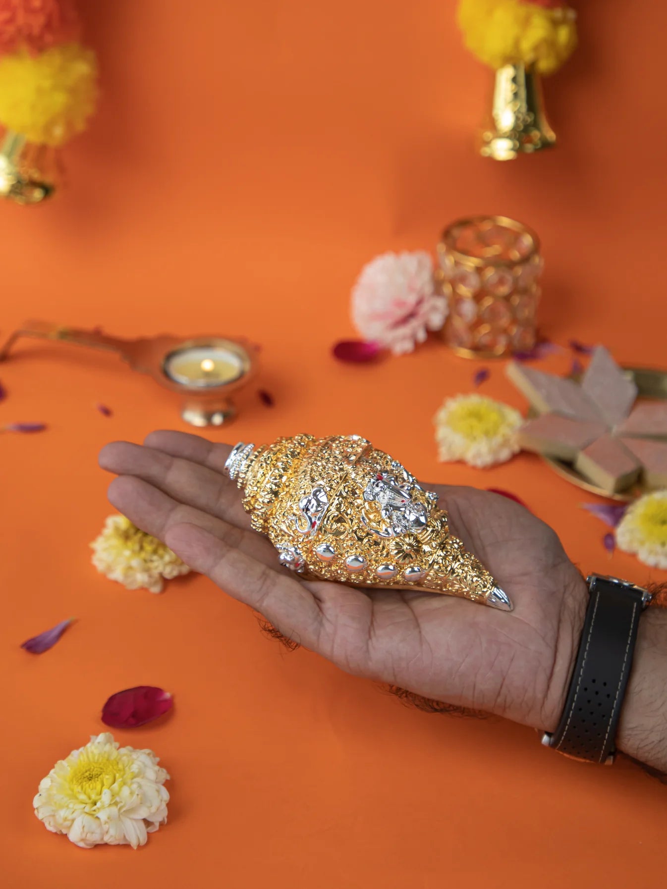 A close-up shot of a person's hand holding a Ganesh Shankh, which is a decorative conch shell with silver and gold plating and intricate carvings. The shankh is a sacred symbol in Hindu rituals and is believed to represent wealth, prosperity, and good luck. The background is an orange color with scattered flower petals and other decorative items for a puja or religious ceremony.