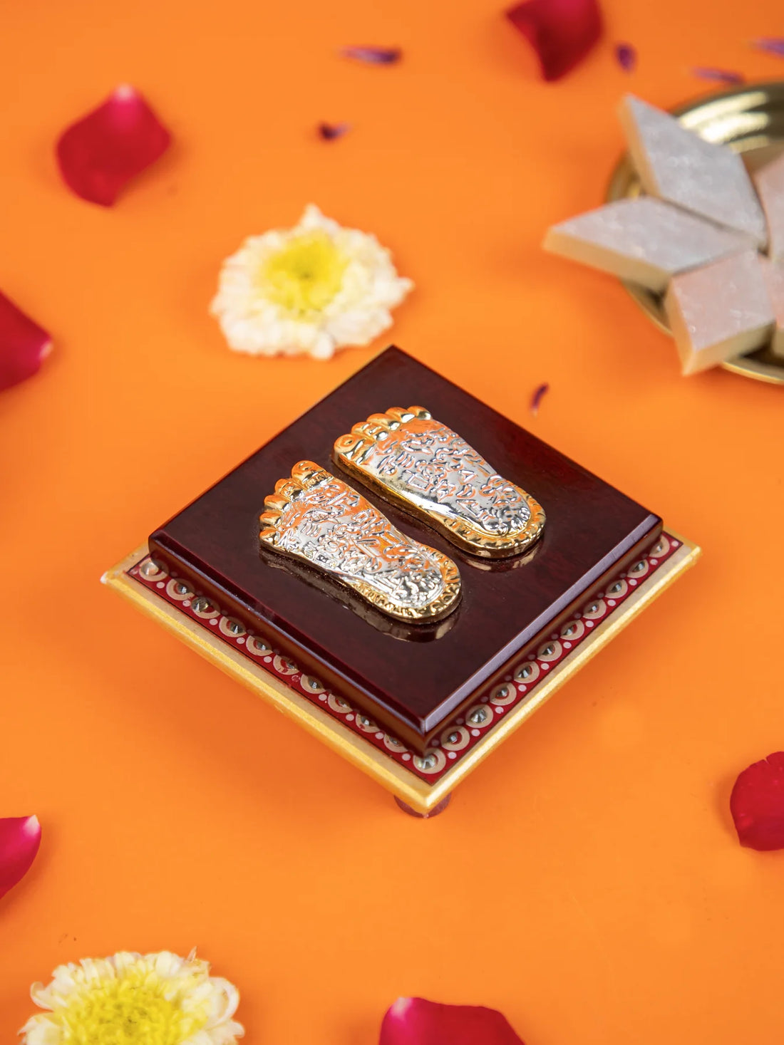 A close-up shot of a decorative item featuring a pair of silver and gold-colored feet impressions, known as "Laxmi Charan Paduka," on a dark wooden base with a golden border. The item is surrounded by rose petals, a white flower, and a few pieces of sweets, likely a type of barfi, on a bright orange surface.