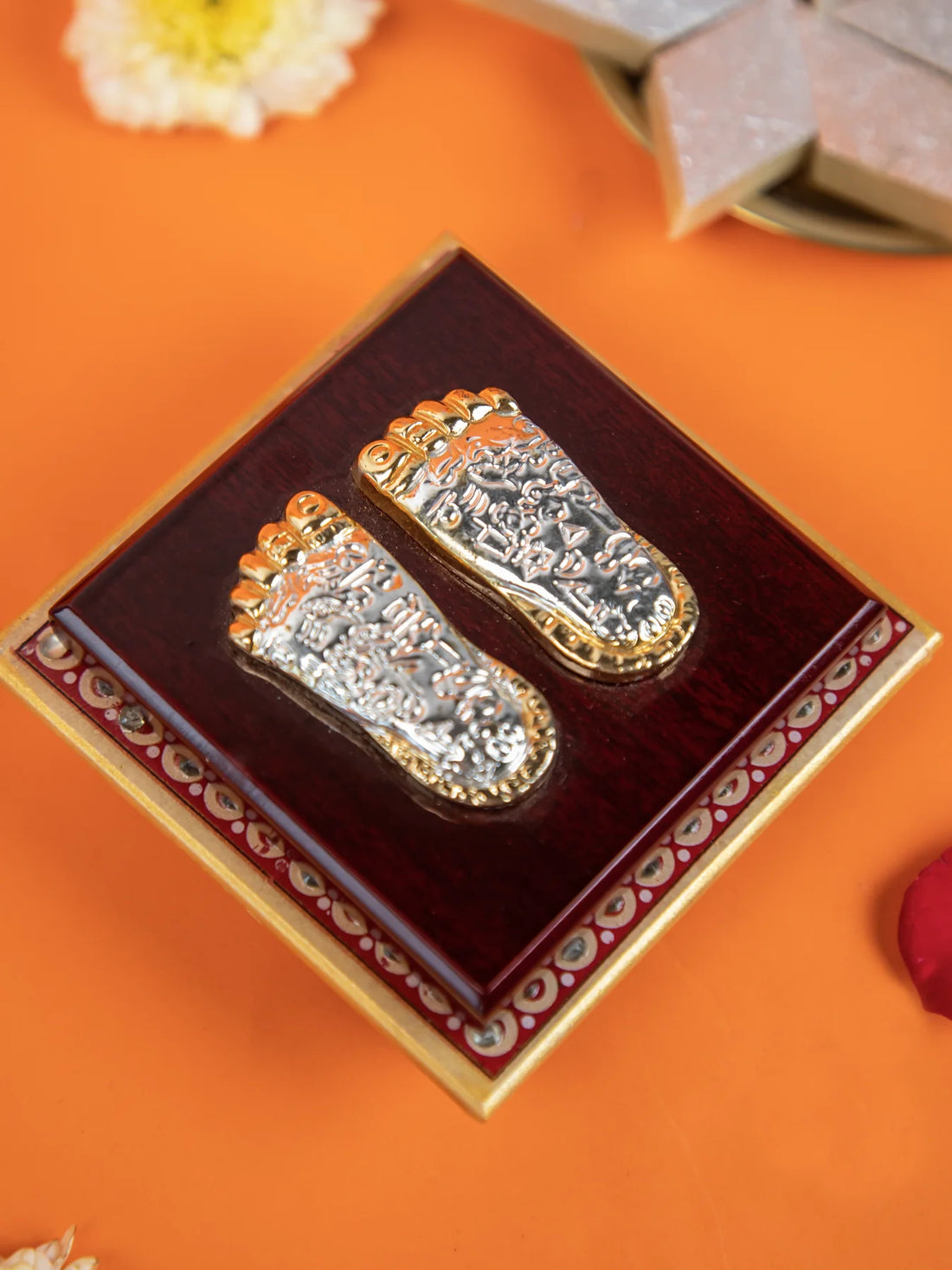 A close-up shot of a decorative item featuring a pair of silver and gold-colored feet impressions, known as "Laxmi Charan Paduka," on a dark wooden base with a golden border. The item is surrounded by rose petals, a white flower, and a few pieces of sweets, likely a type of barfi, on a bright orange surface.