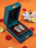 A decorative gift box containing a pair of silver and gold-colored feet, known as Laxmi Charan Paduka, is placed on a dark wooden stand. The open box is lined with a teal velvet fabric, and the entire arrangement sits on an orange surface scattered with red and white flower petals. In the background, there is a plate with sweets.