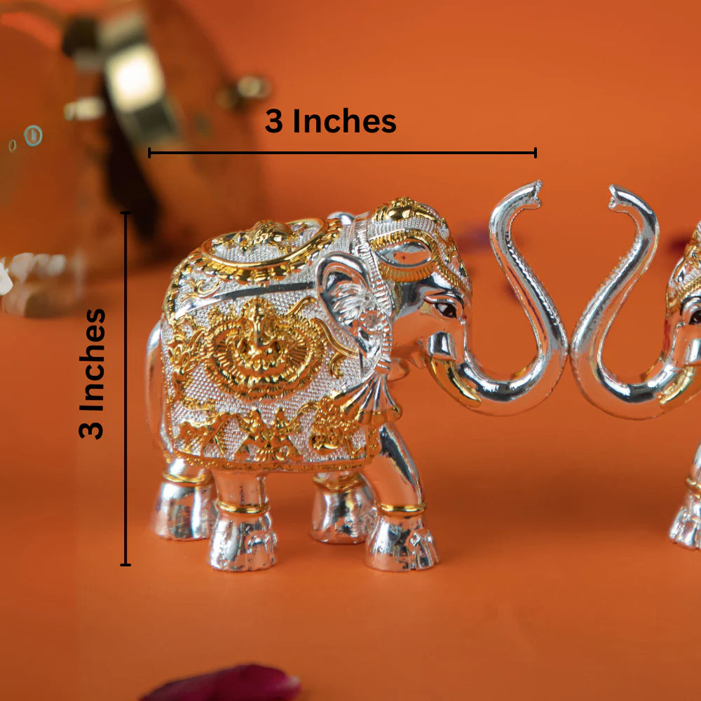 close-up shot of a pair of gold and silver elephant statues with their trunks intertwined. The elephants are intricately decorated with detailed patterns. The image is set against an orange background, with blurred flowers and other decorative items visible in the background.