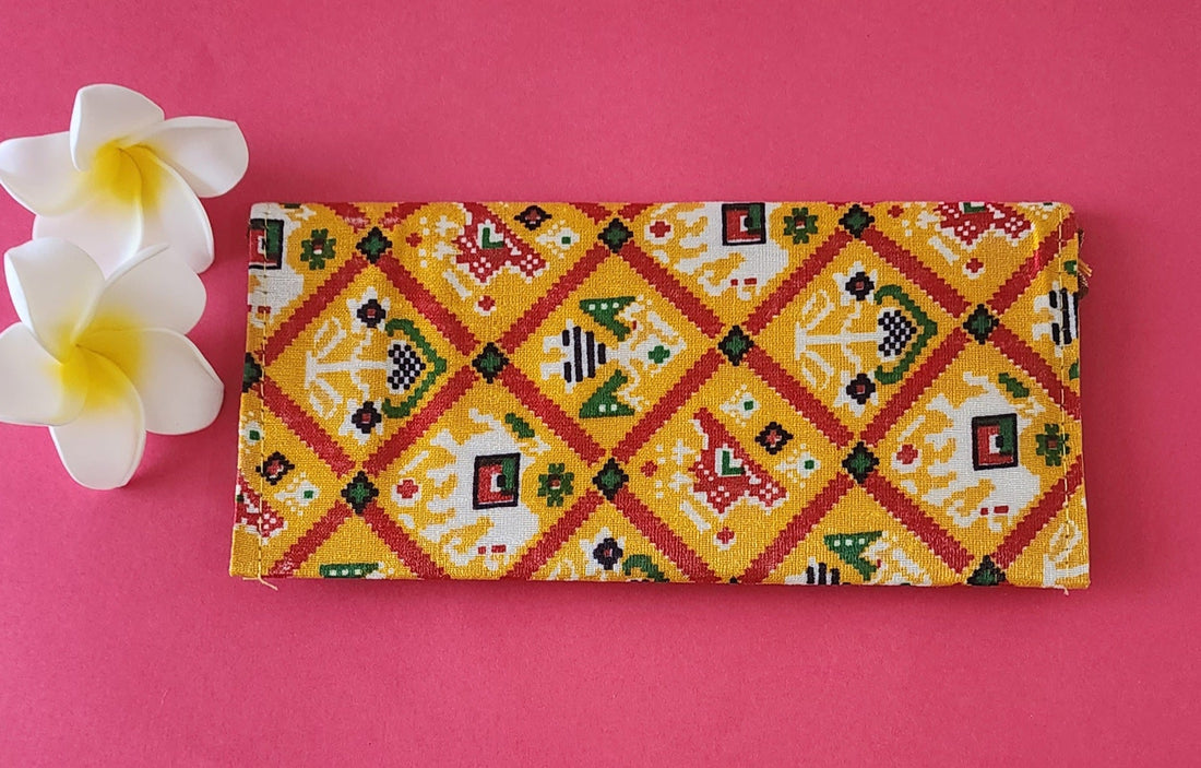A multicolored floral patterned Designer Shagun Envelope placed on a pink surface with two white flowers at the top left corner.