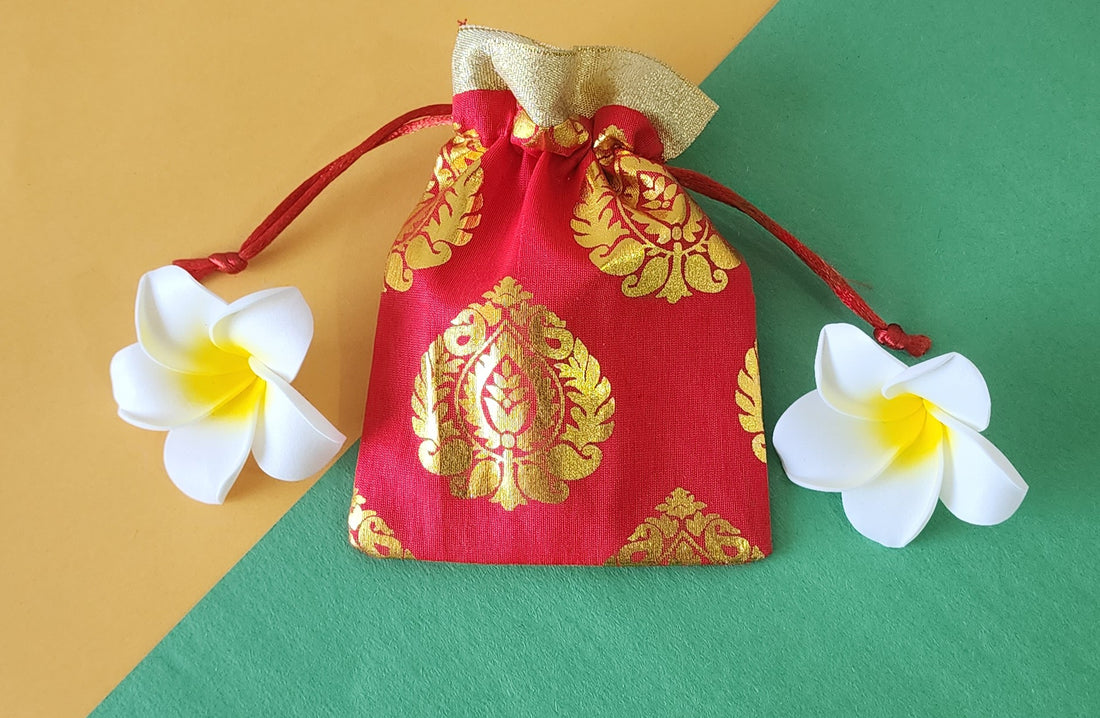 A red and gold patterned fabric potli pouch with a drawstring closure, accompanied by two white flowers on a green background.