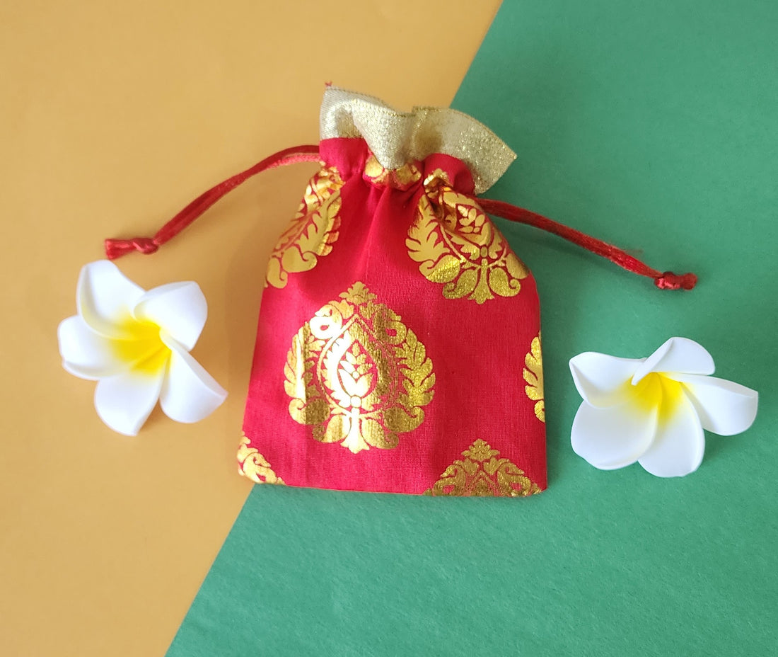 A red and gold patterned fabric potli pouch with a drawstring closure, accompanied by two white flowers on a green background.