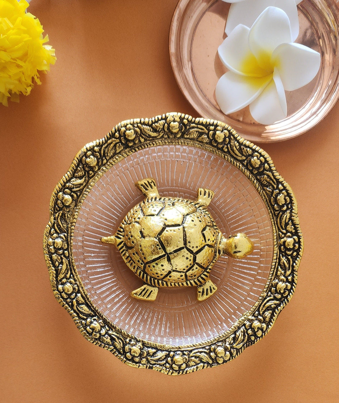 A decorative ceramic bowl in pastel pink color with intricate gold patterns around the rim, containing a small turtle figurine with a detailed, earthy-toned shell.