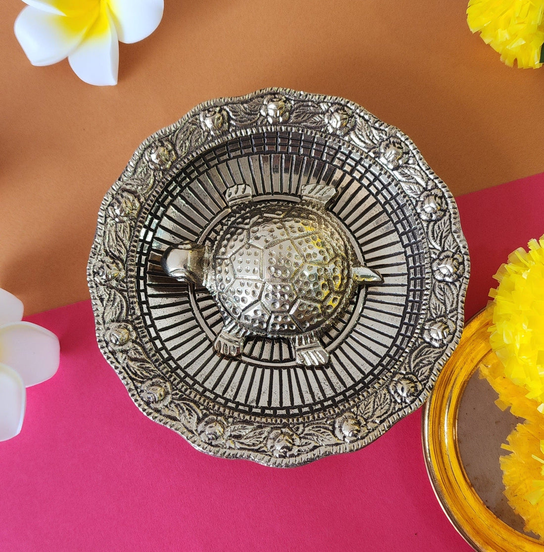 A metal turtle placed on a decorative plate with floral patterns, possibly used for Vaastu Fengshui purposes, displayed against a colorful background.
