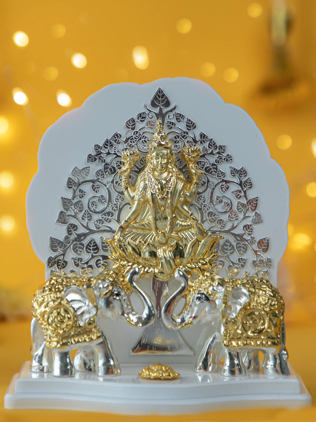 Gajalakshmi idol made of silver and gold-plated material, featuring the goddess Lakshmi seated on a lotus, flanked by two elephants with raised trunks, against a backdrop of a detailed tree design.