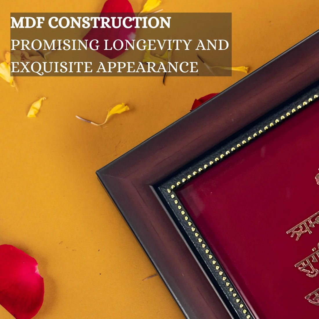 A wooden photo frame with a deep red background and golden text of - Mahamrityunjaya Mantra is placed on a surface. The text inside the frame is in Devanagari script. The frame is set against a yellow wall, with red flower garlands hanging above it. There are also some scattered flower petals and a brass lamp visible on the left side of the image.