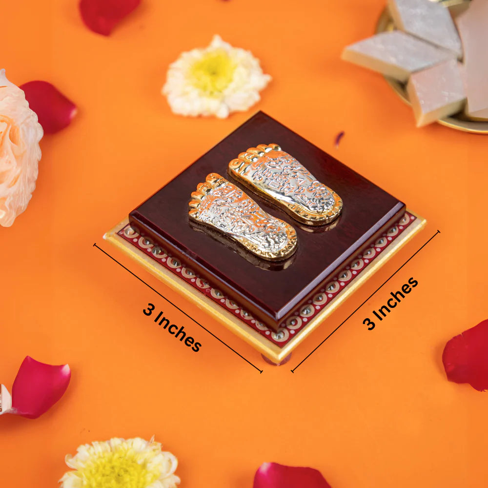 A decorative showpiece of silver-colored Lord Lakshmi Charan Paduka (footprints of the goddess Lakshmi) resting on a square wooden base. The base is 3 inches by 3 inches. The background includes scattered flower petals and traditional Indian sweets.