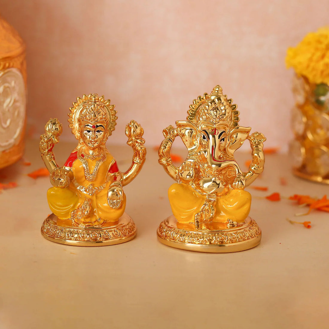 A golden statue of Goddess Lakshmi seated, holding lotuses, next to a golden statue of Lord Ganesha, also seated, with his trunk turned to the left. Both idols are on a reddish-brown surface with a yellow flower garland visible in the background.