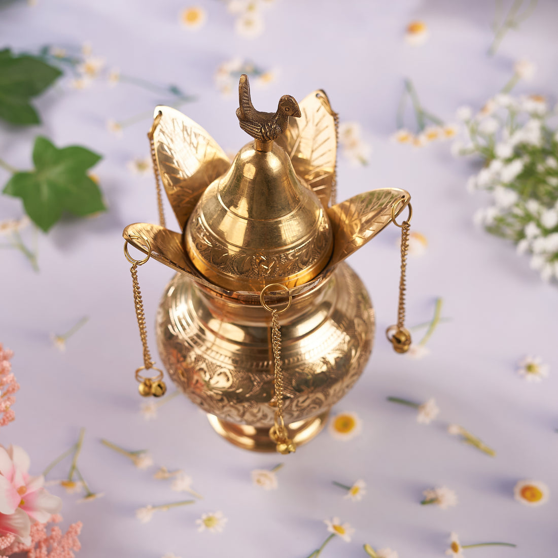 A brass etched handwork lota with intricate designs and a brass coconut, both with a golden shine, set against a floral backdrop.