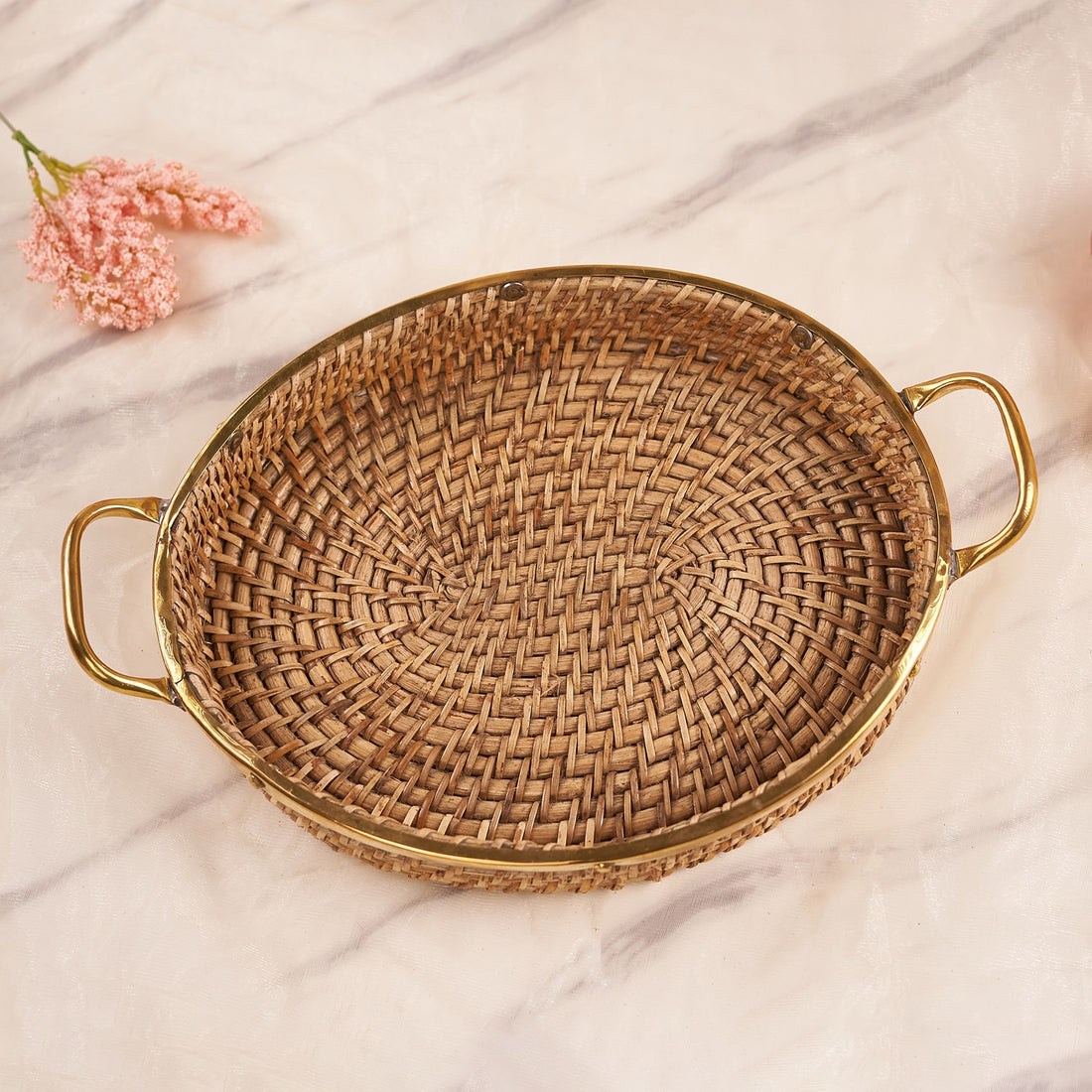 An oval-shaped rattan tray with handles, featuring a beige color and a finely woven design, displayed on a marble surface with a pink flower in the corner.