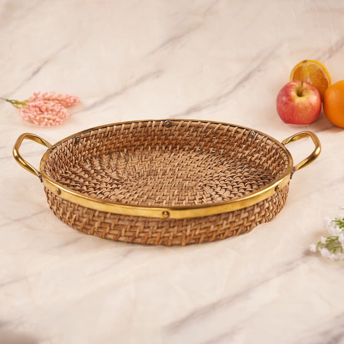 An oval-shaped rattan tray with handles, featuring a beige color and a finely woven design, displayed on a marble surface with a pink flower in the corner.