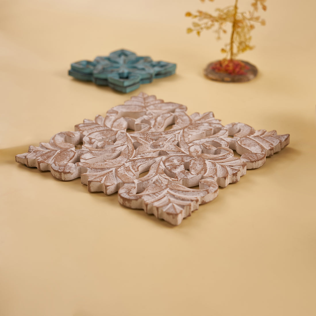 A wooden carved trivet with an antique white finish, featuring intricate floral patterns, displayed on a yellow background.
