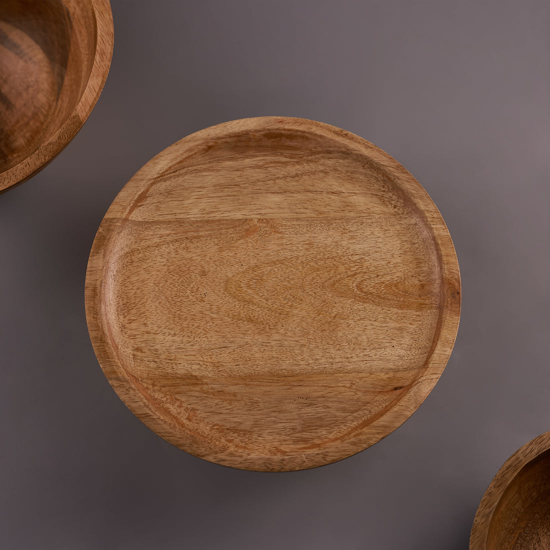 A mango wood cake stand displaying a cake, with a smaller stand visible in the background and a potted plant to the side.