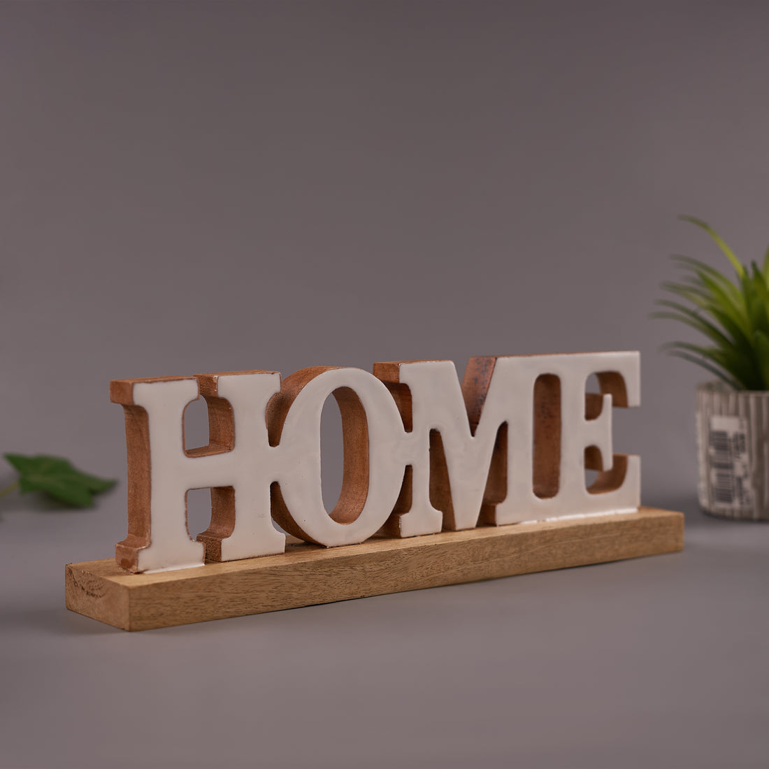 A wooden 'HOME' word sign with a natural finish, displayed on a gray surface with a potted plant and decorative flower nearby.