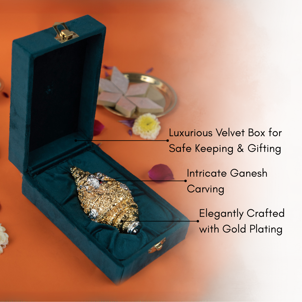 A golden and silver conch shell, or shankh, with intricate carvings rests on a vibrant orange surface, surrounded by scattered pink and yellow flower petals. In the background, there is a golden decorative object and some sweets.
