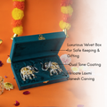 A luxurious velvet box, open to reveal two decorative elephant figurines. The figurines have a dual-tone coating and intricate carvings of what appear to be Laxmi and Ganesh. The box is shown on a background of orange with floral garlands.