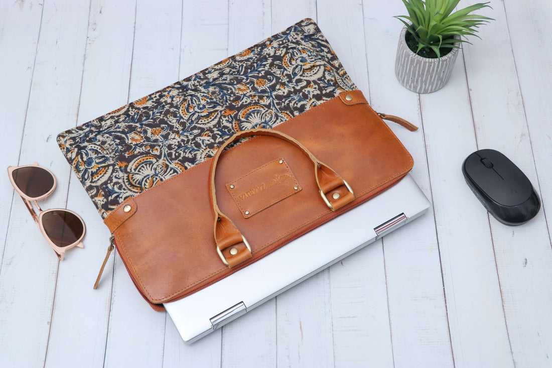 A dark blue leather laptop sleeve with block print design, displayed on a wooden surface alongside a laptop, sunglasses, and a houseplant.
