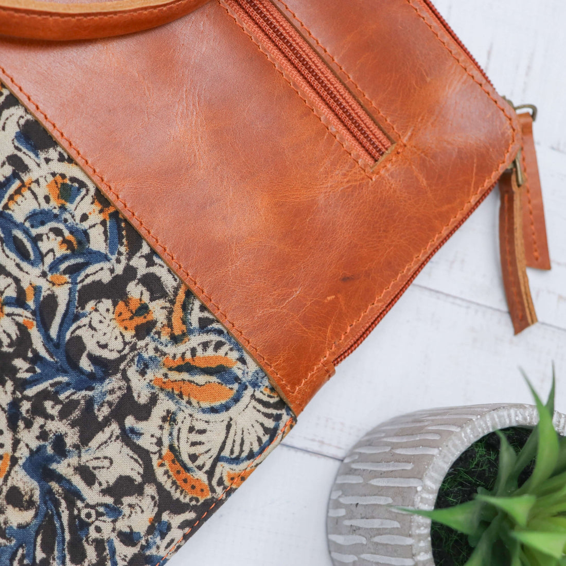A dark blue leather laptop sleeve with block print design, displayed on a wooden surface alongside a laptop, sunglasses, and a houseplant.