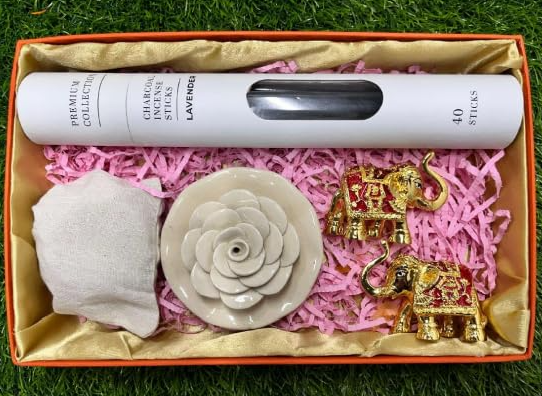A gift set containing incense sticks, a ceramic rose incense holder, two red elephant figurines, and dry fruit pouch, all presented in an open gift box with a pink interior lining.