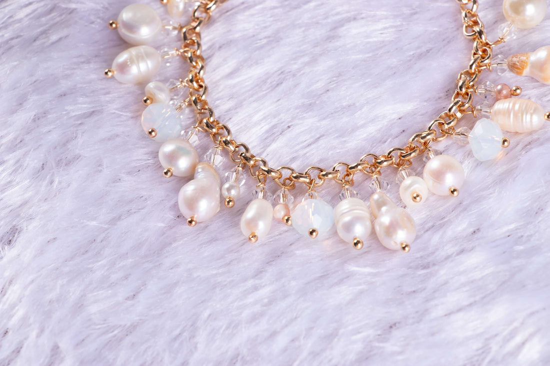 A bracelet featuring a series of round baroque pearl charms linked together with gold-colored accents, displayed on a white furry surface with a floral arrangement in the background.