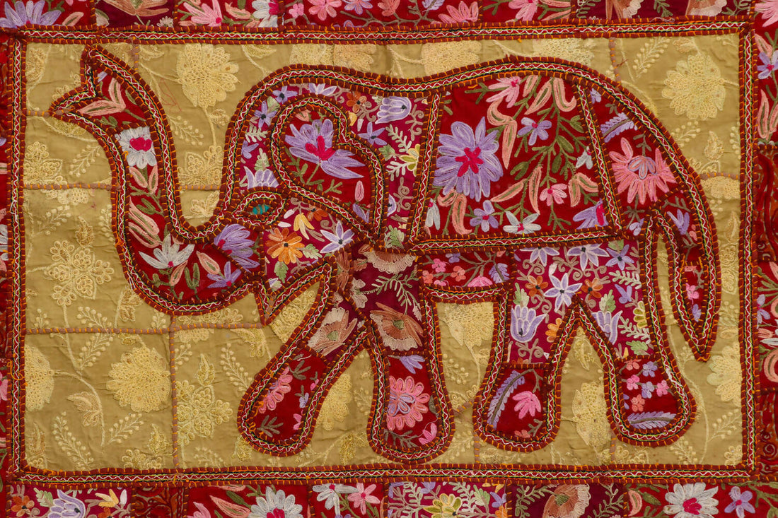 A wall decor featuring an embroidered elephant design with intricate patterns, predominantly in pink and red colors, on a gold background with a red border.