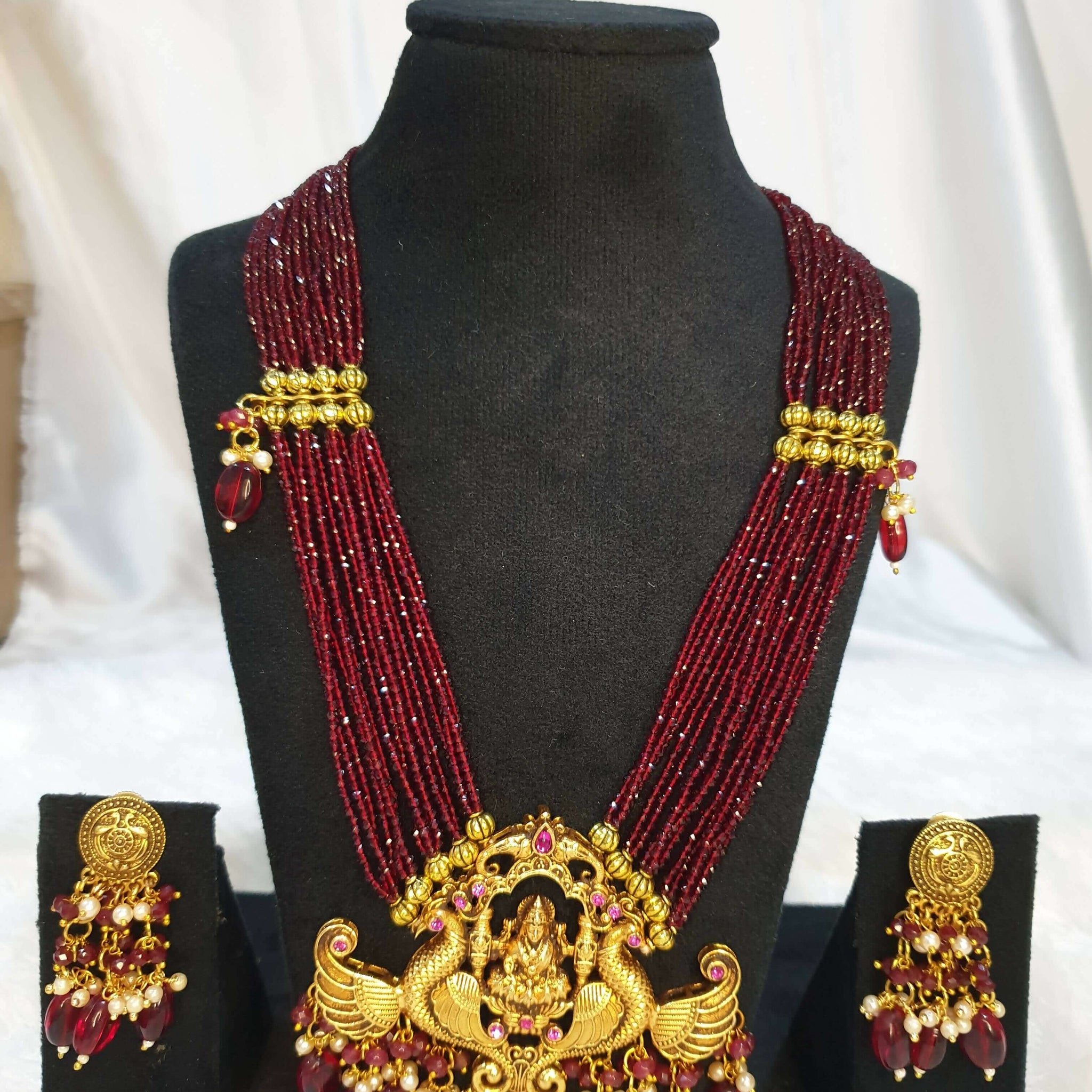A multi-layered maroon necklace with gold detailing and a gold pendant, displayed on a mannequin with matching earrings.